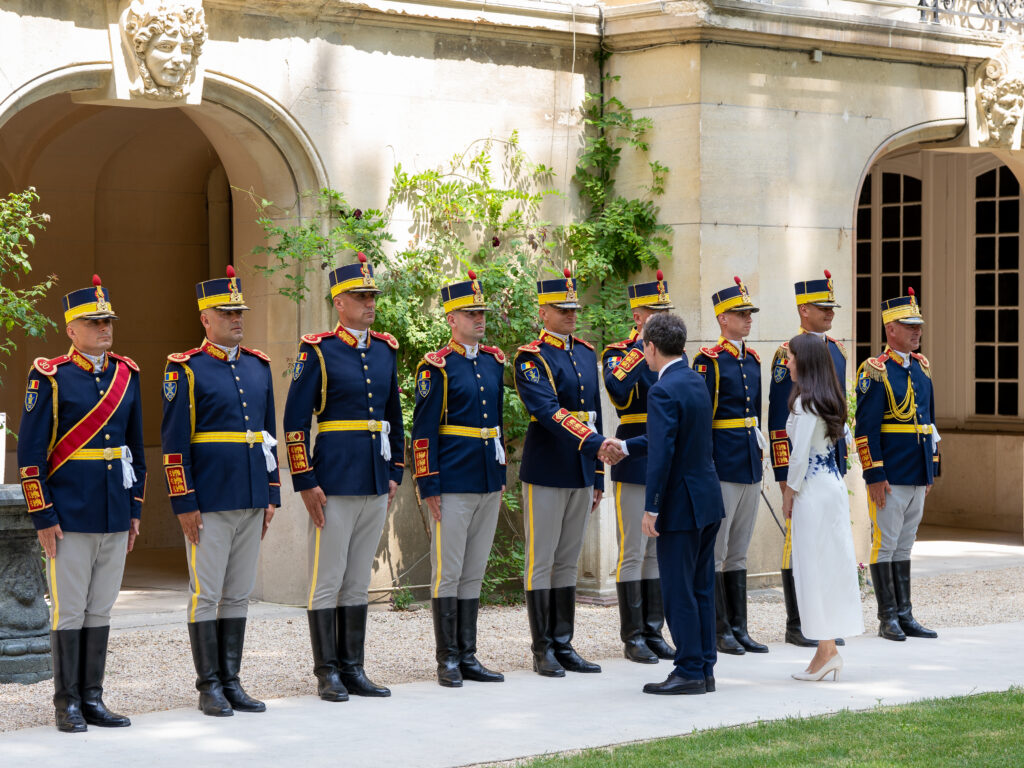 Line of uniformed guards in blue and gold dress uniforms standing outdoors, shaking hands with a man in a navy suit and a woman in a white dress, with a stone building and greenery behind.