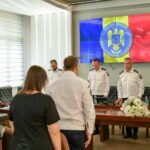 Group of people in a formal meeting room; three uniformed officers stand at a long wooden table with floral arrangements while attendees sit nearby, a large screen shows the Romanian Police emblem with blue, yellow, and red panels in the background.