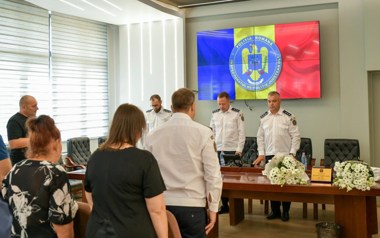 Group of people in a formal meeting room; three uniformed officers stand at a long wooden table with floral arrangements while attendees sit nearby, a large screen shows the Romanian Police emblem with blue, yellow, and red panels in the background.