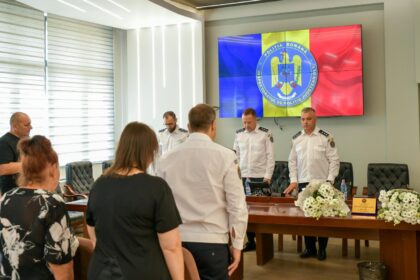 Group of people in a formal meeting room; three uniformed officers stand at a long wooden table with floral arrangements while attendees sit nearby, a large screen shows the Romanian Police emblem with blue, yellow, and red panels in the background.