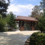 Wooden gazebo with a tiled roof in a green garden, on a sunny day with a paved foreground.