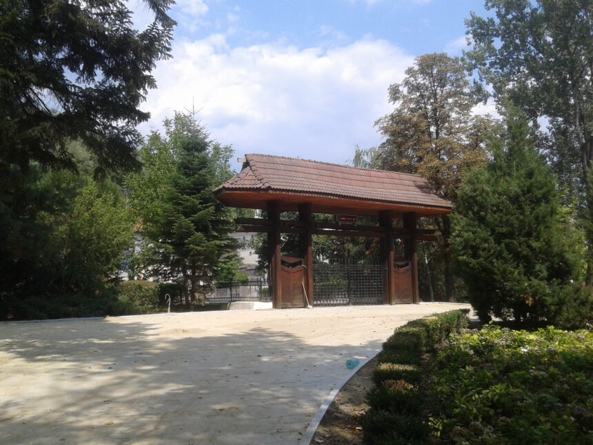 Wooden gazebo with a tiled roof in a green garden, on a sunny day with a paved foreground.