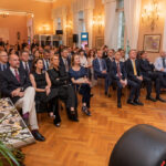 Audience of formally dressed men and women seated in a chandelier-lit room during a conference or ceremony.