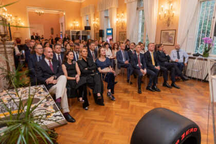 Audience of formally dressed men and women seated in a chandelier-lit room during a conference or ceremony.