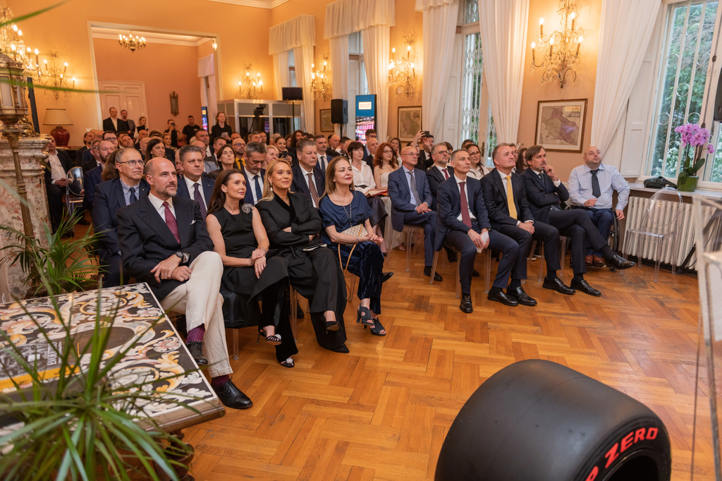 Audience of formally dressed men and women seated in a chandelier-lit room during a conference or ceremony.