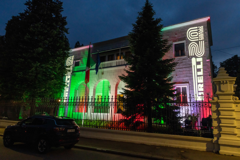 Night view of a historic stone building lit in green, white, and red lights behind a wrought-iron fence; a dark SUV parked on the street.