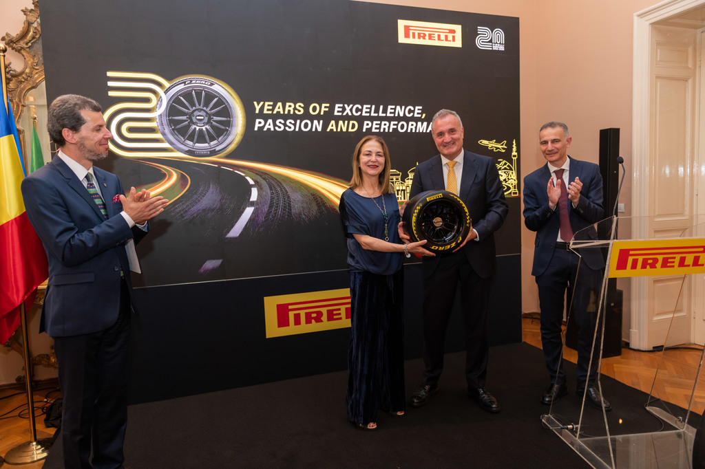 Group of four adults on a stage at a Pirelli event, with a large backdrop reading'20 Years of Excellence, Passion and Performance' and a wheel display being handed to a man in a suit.