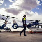 Police officer in uniform walking on a helicopter pad with blue-and-white government helicopters in the background.