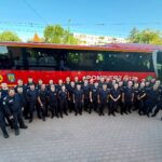 Group of uniformed firefighters posing in front of a red rescue bus labeled 'POMPIERII' on a sunny day.