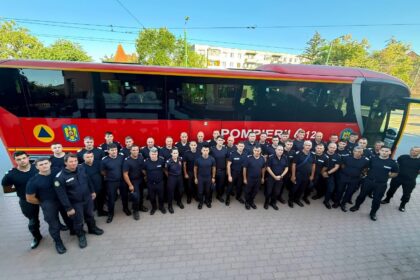 Group of uniformed firefighters posing in front of a red rescue bus labeled 'POMPIERII' on a sunny day.