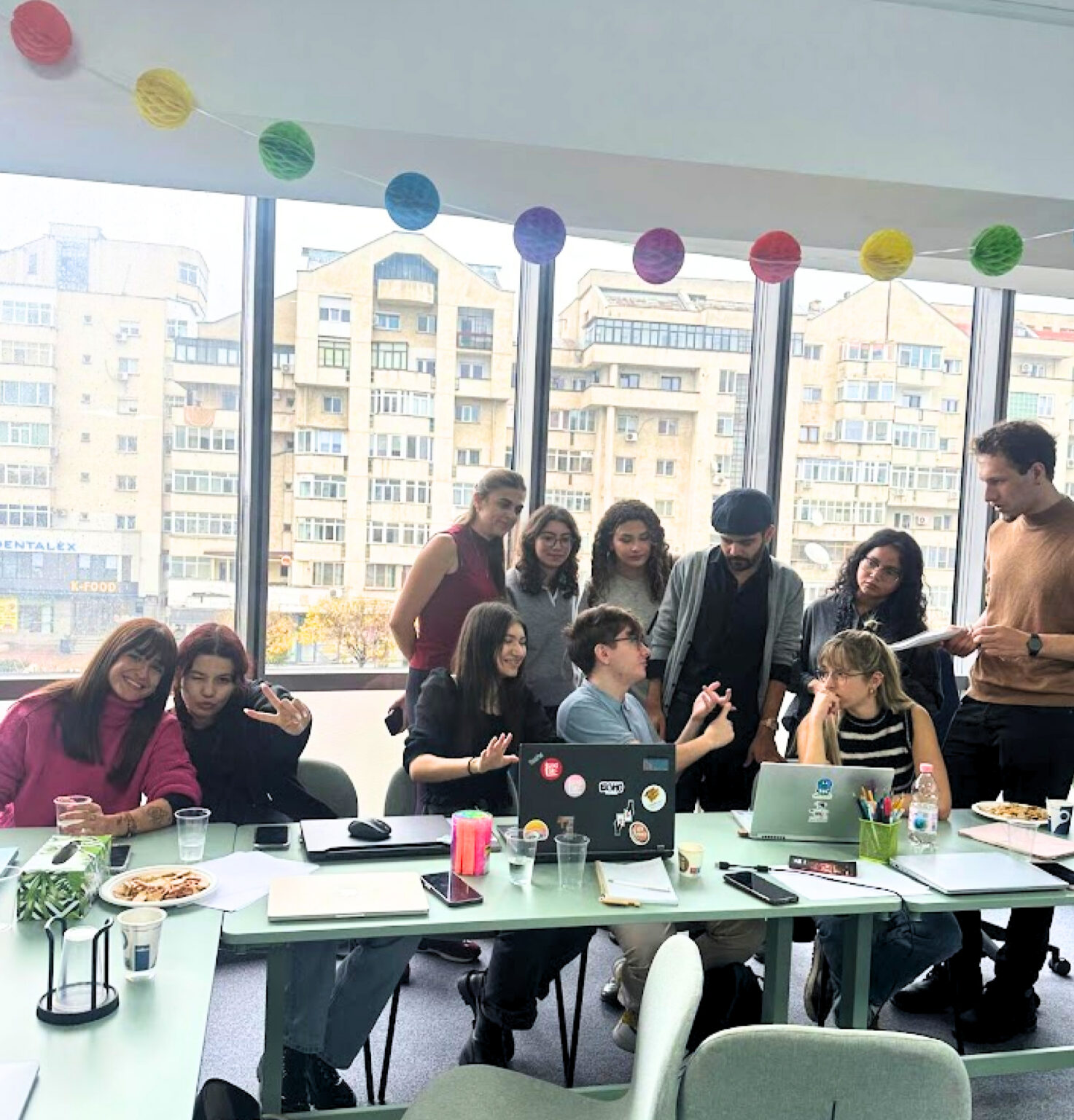 Group of people around a glass table with laptops, snacks, and drinks in a bright room, a casual gathering or meeting.