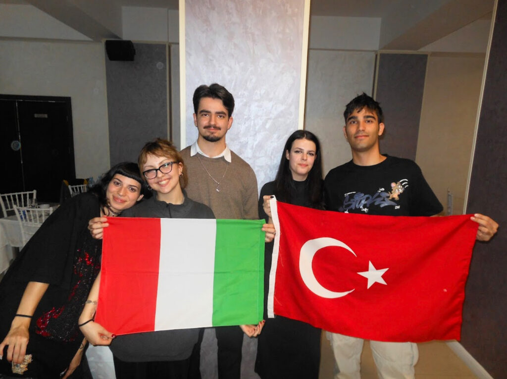 Five young adults pose indoors, holding Italian (green, white, red) and Turkish flags and smiling at the camera.