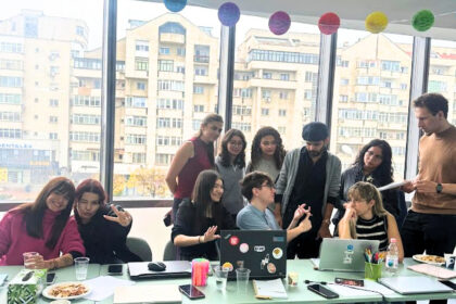 Group of people around a glass table with laptops, snacks, and drinks in a bright room, a casual gathering or meeting.