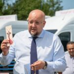 Bald man in a blue shirt and tie holding a packaged meat skewer outdoors, with a white van in the background.
