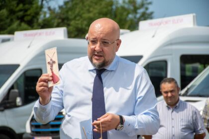 Bald man in a blue shirt and tie holding a packaged meat skewer outdoors, with a white van in the background.