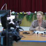 Woman in a leopard-print blouse sits at a table with multiple microphones while a video camera films her interview.