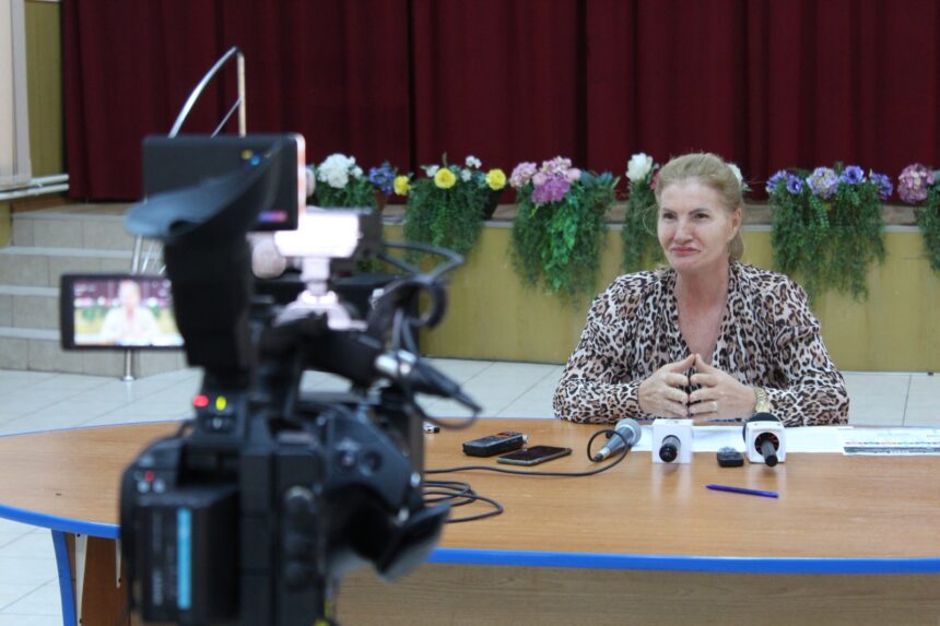 Woman in a leopard-print blouse sits at a table with multiple microphones while a video camera films her interview.