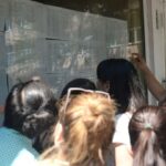 Group of people gathered around a glass bulletin board, reading posted schedules outside a building with greenery nearby.