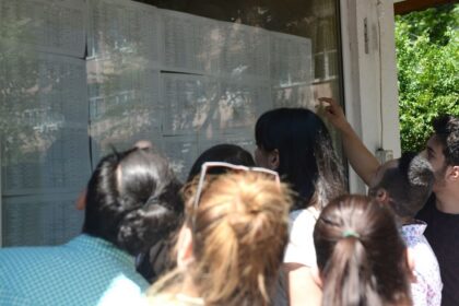 Group of people gathered around a glass bulletin board, reading posted schedules outside a building with greenery nearby.