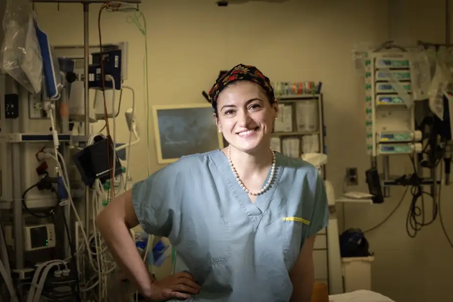 sinziana dumitra - Jurnalul Olteniei – Știri din Craiova și Oltenia Female nurse in blue scrubs smiling in a hospital room with medical equipment in the background.