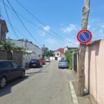A residential street with parked cars on both sides and a blue no-parking sign on a wooden utility pole against a clear blue sky.