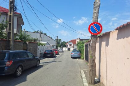 A residential street with parked cars on both sides and a blue no-parking sign on a wooden utility pole against a clear blue sky.