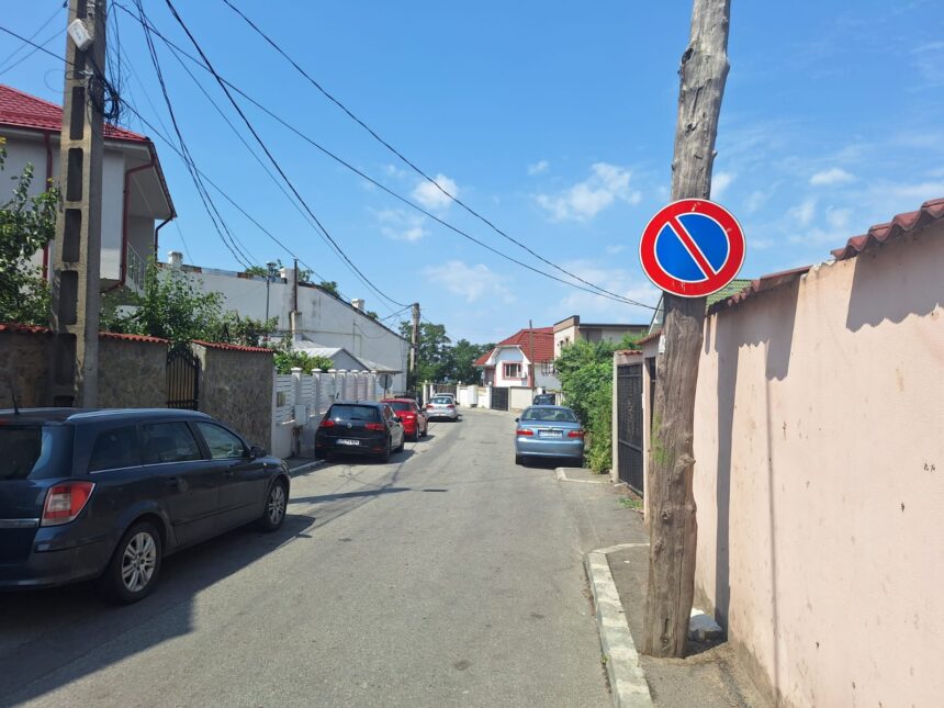 A residential street with parked cars on both sides and a blue no-parking sign on a wooden utility pole against a clear blue sky.