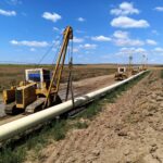Oil field with yellow drilling rigs laying pipeline across muddy ground under a blue sky with scattered clouds.
