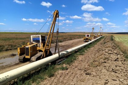 Oil field with yellow drilling rigs laying pipeline across muddy ground under a blue sky with scattered clouds.