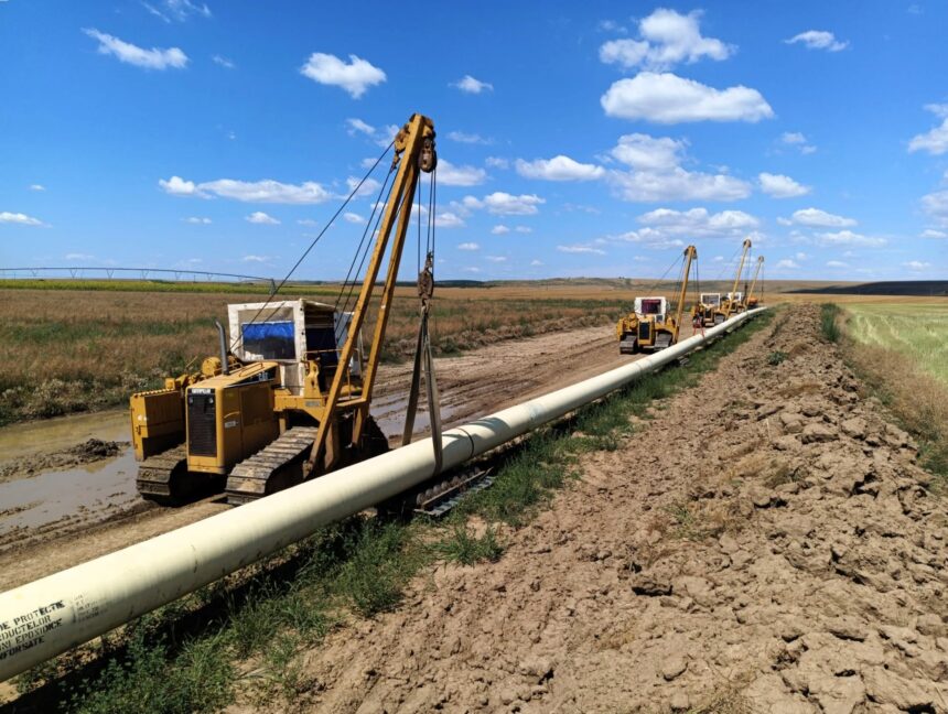 Oil field with yellow drilling rigs laying pipeline across muddy ground under a blue sky with scattered clouds.