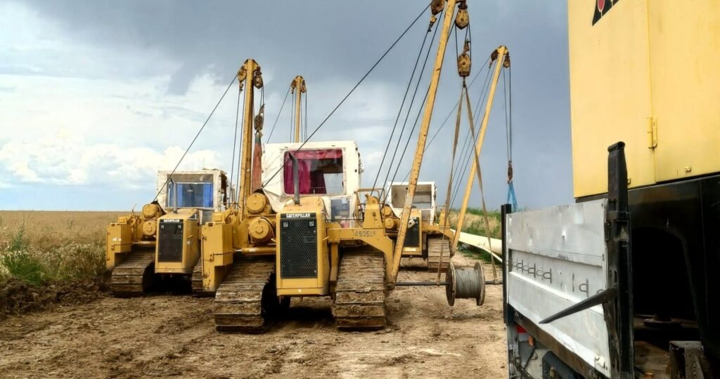 subtraversare raul olt 2 - Jurnalul Olteniei – Știri din Craiova și Oltenia Group of yellow Caterpillar crawler tractors with booms and cables on a dirt field, under a cloudy sky.