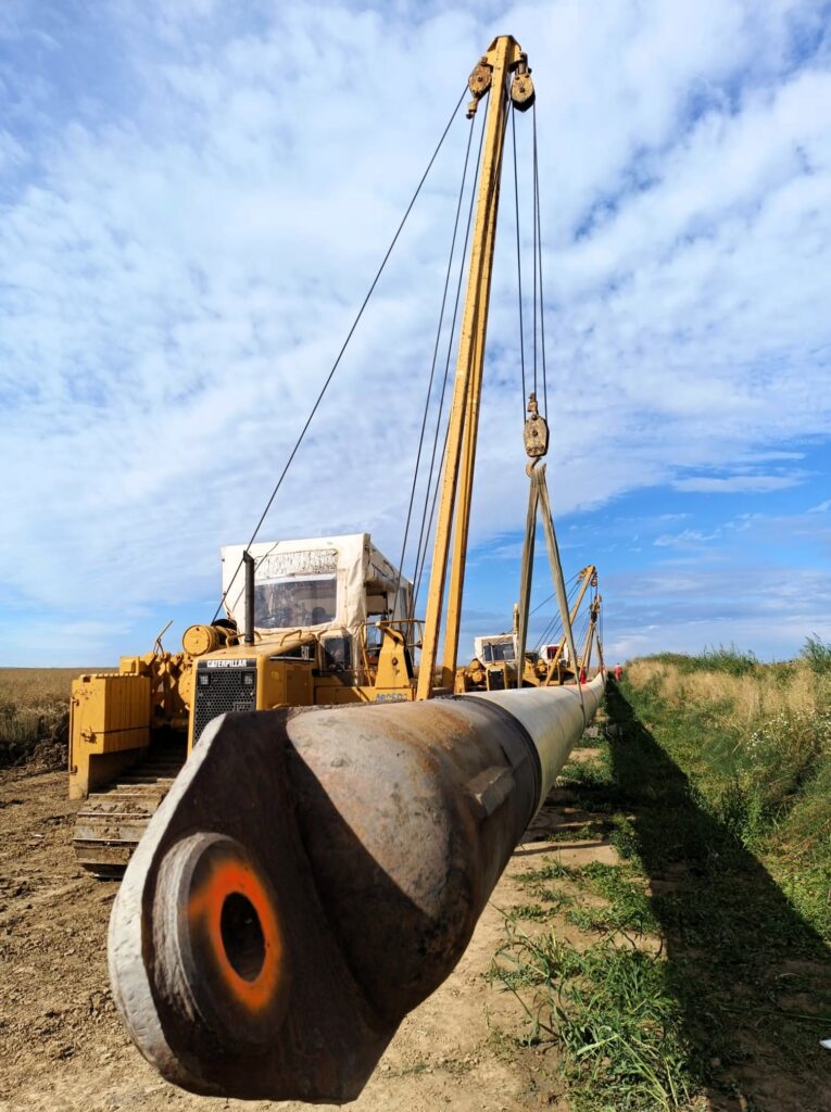 subtraversare raul olt - Jurnalul Olteniei – Știri din Craiova și Oltenia Large yellow construction crane and piping equipment on a rural pipeline project under a blue sky with scattered clouds, cables visible overhead to lift a heavy pipe