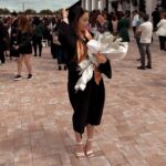 Graduate in a black gown adjusts her cap while holding a large bouquet wrapped in white paper outdoors at a graduation ceremony.