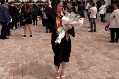 Graduate in a black gown adjusts her cap while holding a large bouquet wrapped in white paper outdoors at a graduation ceremony.