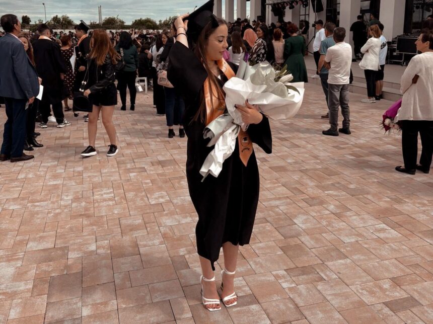 Graduate in a black gown adjusts her cap while holding a large bouquet wrapped in white paper outdoors at a graduation ceremony.