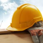 Yellow hard hat and work gloves resting on a wooden beam at a construction site, with cranes and a city skyline in the background.