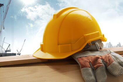Yellow hard hat and work gloves resting on a wooden beam at a construction site, with cranes and a city skyline in the background.