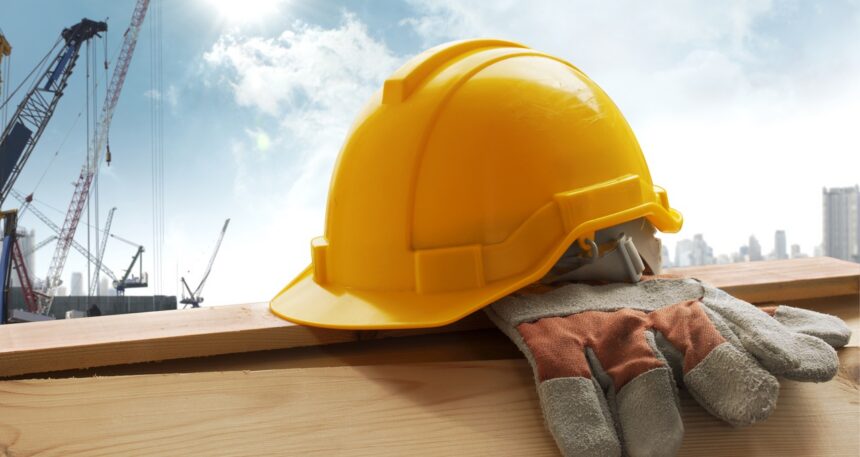 Yellow hard hat and work gloves resting on a wooden beam at a construction site, with cranes and a city skyline in the background.