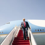 Man in a dark suit and red tie giving a thumbs-up while stepping down from a large blue airplane on a tarmac.