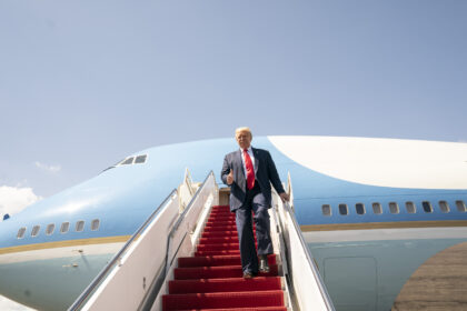 Man in a dark suit and red tie giving a thumbs-up while stepping down from a large blue airplane on a tarmac.