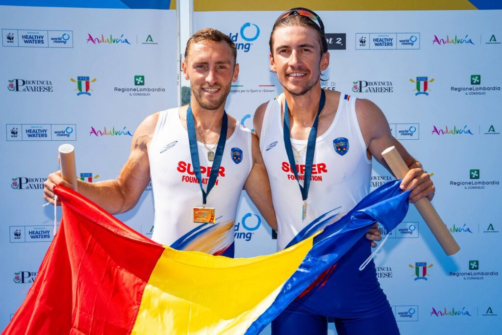 Two smiling male rowers with medals around their necks pose in white singlets, holding a large red-yellow-blue flag against a sponsor backdrop.