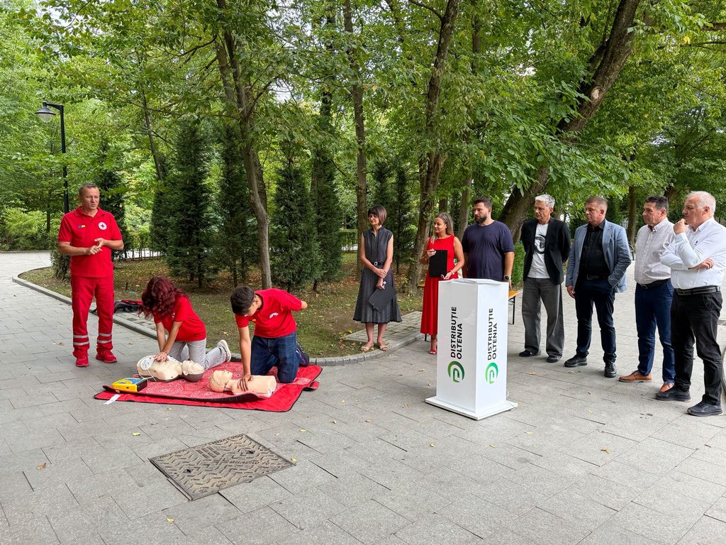 CPR training demonstration outdoors with mannequins on red mats and a presenter in a red uniform addressing onlookers under trees.
