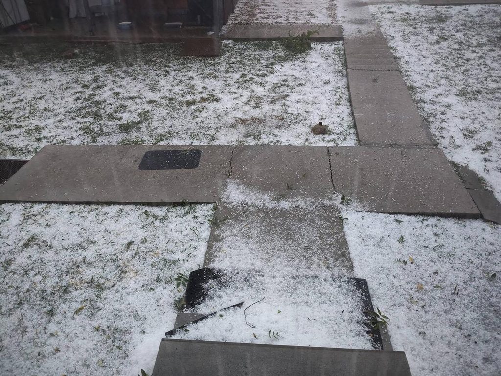Outdoor paved walkway made of square concrete slabs with a light layer of hail across the ground and grass patches visible beneath.