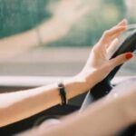 Close-up of a driver's hands on the steering wheel, with red nail polish and a bracelet visible in a car.