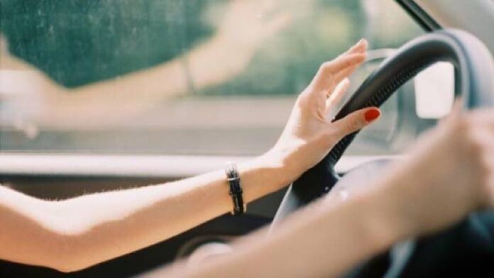Close-up of a driver's hands on the steering wheel, with red nail polish and a bracelet visible in a car.