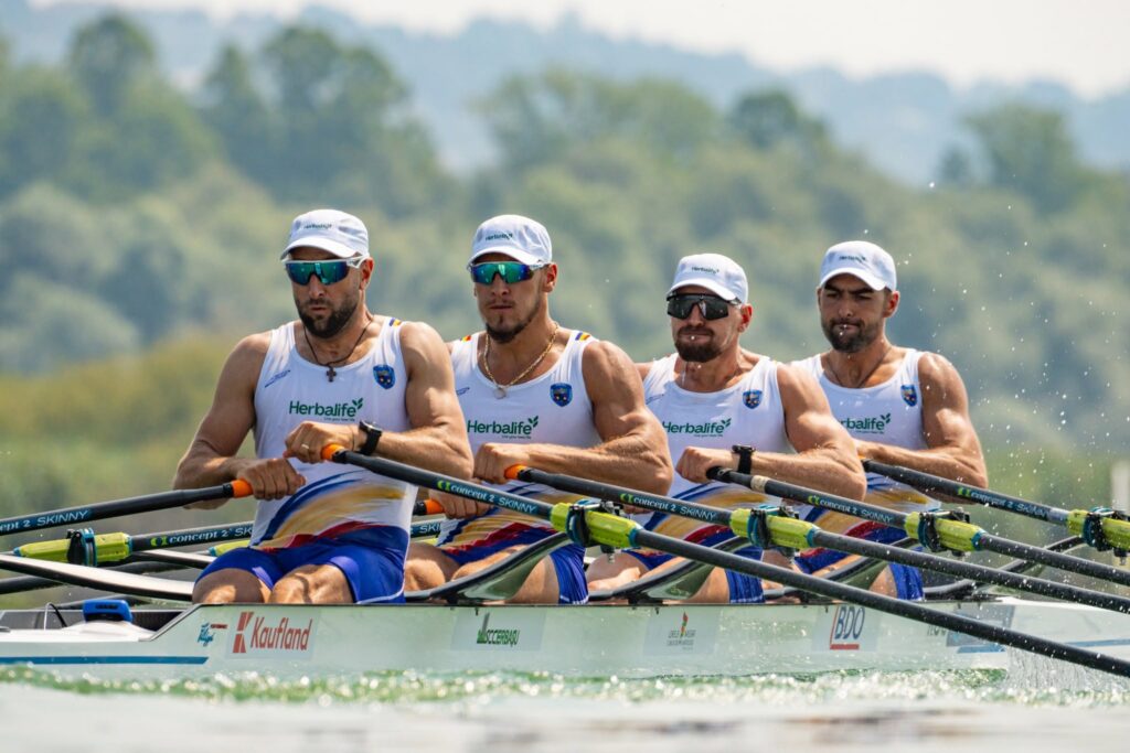 Four male rowers in white Herbalife caps rowing a racing shell on a river, synchronized with oars in action behind a low boat.