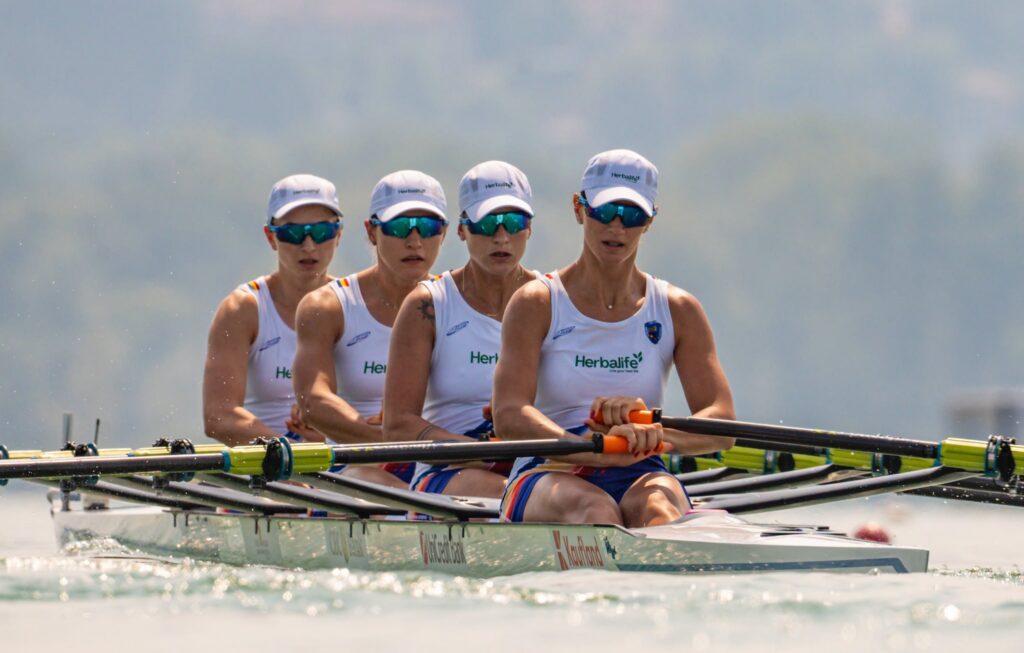 Four female rowers in white uniforms paddle in unison on a racing shell, wearing white caps and blue mirrored sunglasses, with oars in the water and a misty lake backdrop.