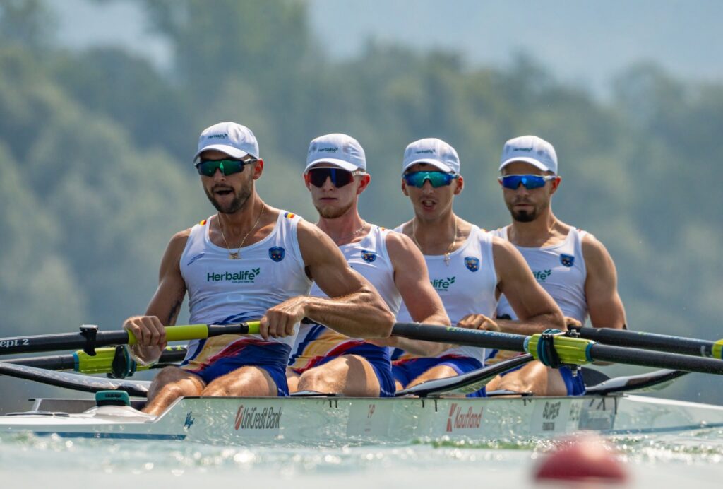 Four rowers in a racing shell paddling in sync, wearing matching white uniforms and caps, with oars in foreground.