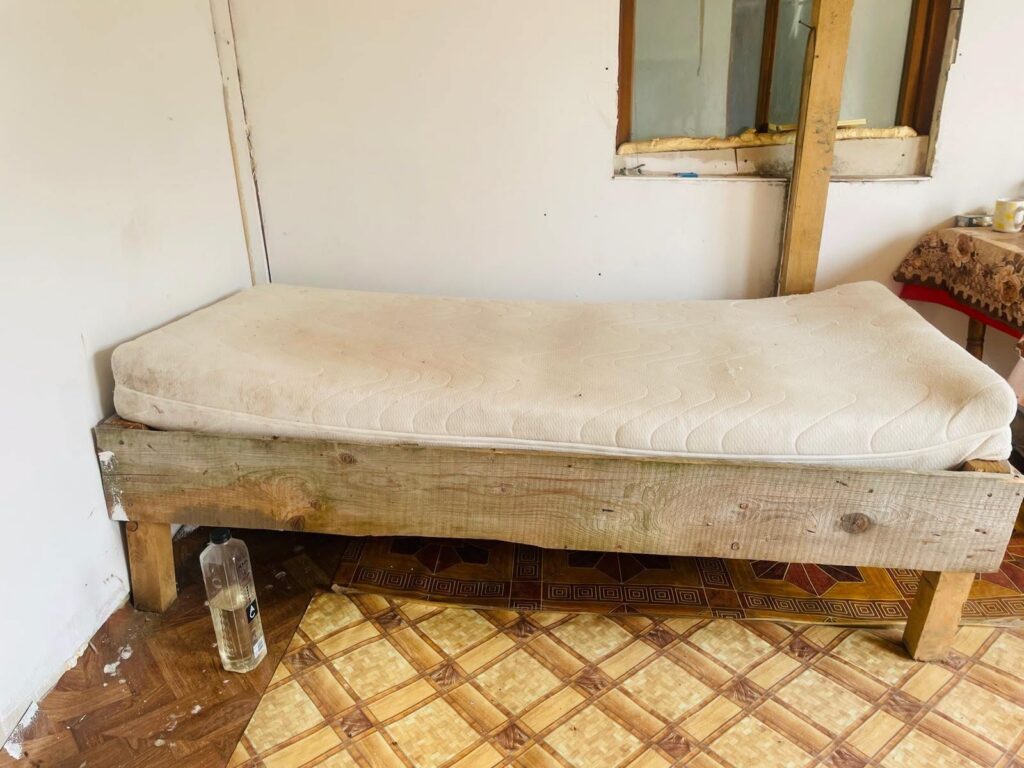 A simple wooden bed frame with a white mattress in a bare, unfinished room; a plastic water bottle sits on the floor beside the bed.