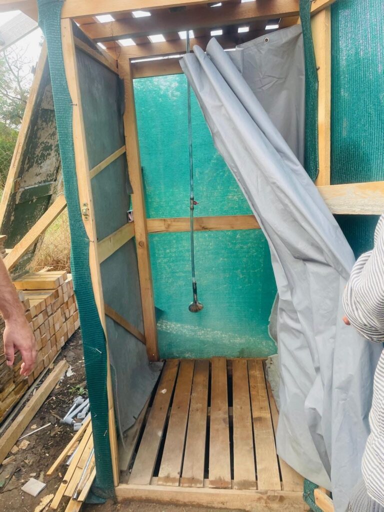 DIY outdoor shower stall under construction with green mesh walls, wooden frame, and a hanging shower head inside.